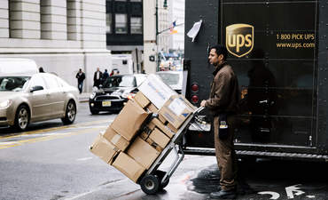 在纽约，联合包裹快递公司的快递员正站在他的卡车旁等着过马路，他手里拿着一辆装满包裹的手推车