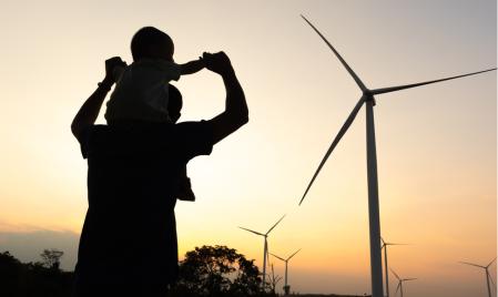 将孩子抱在肩上的人的轮廓，在日落时的距离中可以看到风力涡轮机。