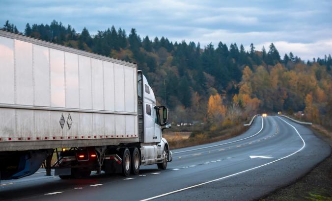 White long haul semi truck with semi trailer running with turned on headlights on rain dusted wet raining highway driving with another cars traffic in twilight evening time
