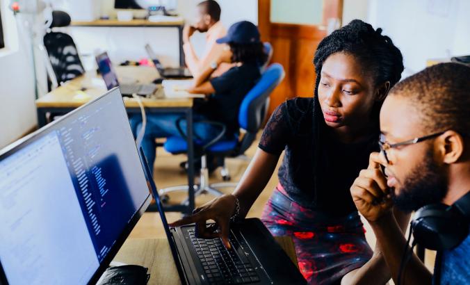 Two people working on computers. There are additional people in a blurred background.